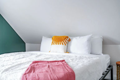 Cozy attic/loft bedroom with sloped ceiling, white bed and fluffy duvet, mustard tassel accent pillow, pink folded throw at the foot and a green accent wall.