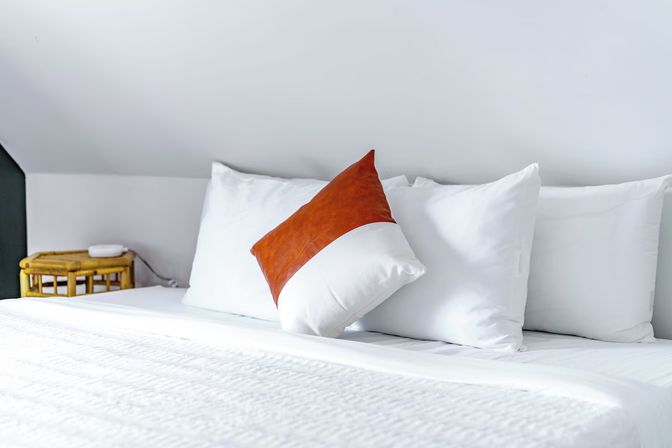 Bright minimalist bedroom with crisp white linens, three plump pillows and a rust-orange accent cushion on a neatly made bed beside a small rattan nightstand