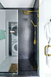Sleek modern walk-in shower with black herringbone tile, matte gold rain showerhead and handheld, frameless glass door with gold handle; stacked washer-dryer visible through doorway and white robe on hook