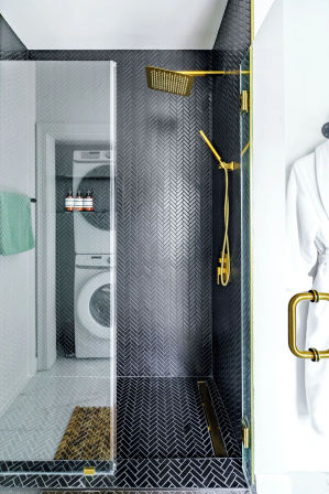 Sleek modern walk-in shower with black herringbone tile, matte gold rain showerhead and handheld, frameless glass door with gold handle; stacked washer-dryer visible through doorway and white robe on hook