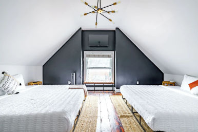 Bright loft attic bedroom with twin beds and white bedding, dark triangular accent wall framing a central window, modern sputnik chandelier and reclaimed wood floors.