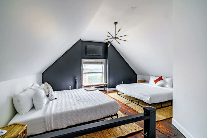 Cozy attic loft bedroom with two white-dressed beds, angled white ceiling and black accent wall with window and wall-mounted TV, mid-century chandelier, hardwood floors and woven rugs — bright, modern rustic vibe.