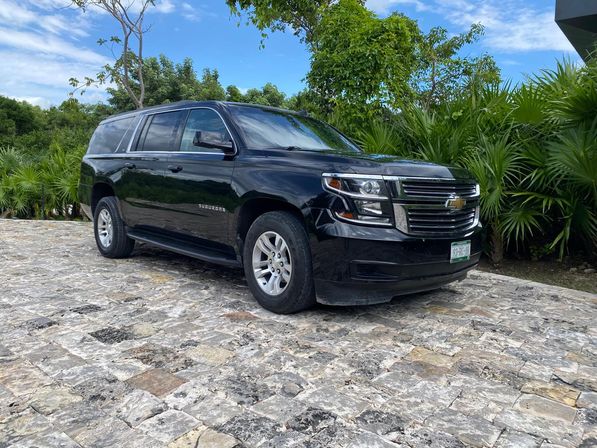 Black Chevrolet Suburban SUV parked on a stone-paved driveway framed by tropical palms under a bright blue sky.