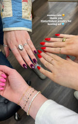 Four hands displaying fresh manicures—hot pink, deep plum and bright red almond-shaped nails—paired with delicate gold rings and layered bracelets over a wood floor, salon-style group manicure.