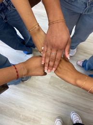 Four hands stacked in a circle showing gold bracelets, rings and French manicure, casual denim and sneakers on a light wood floor