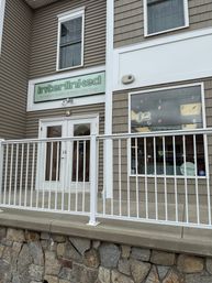 Charming jewelry boutique storefront with mint-green sign, glass double doors and neon "Open" window sign, beige siding, white metal railing and stone foundation.