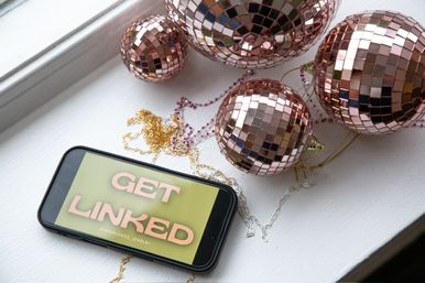 Smartphone reading 'GET LINKED' beside rose-gold mirrored disco ball ornaments and tangled gold and silver chains on a white windowsill