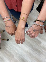 Three wrists showing trendy stacked bracelets—colorful flower-bead strand, delicate gold chain with clover charm, and mixed stone and silver bead bracelets—over a light wood floor with casual shoes visible.