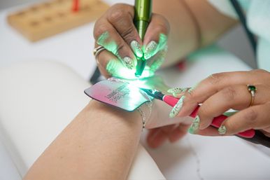 Close-up of hands with decorative nails using a handheld green laser pen and pliers to work on a metal tag and chain bracelet on a wrist