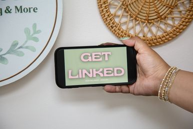 Hand wearing gold beaded bracelets holds a smartphone horizontally displaying pink "GET LINKED" text on a green background, set against a white wall with woven rattan decor and a leafy sign.