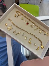 Close-up jewelry flatlay of a gold chain and assorted dainty gold charms (sunflower, key, heart, boot, moon, cherry, initial pendant) neatly arranged on a beige velvet jewelry tray on a glass table