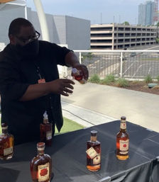 Masked server pours amber whiskey into a glass at an outdoor rooftop tasting table with several bottles on a black tablecloth and an urban parking garage and skyline in the background.