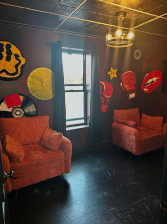 Cozy eclectic lounge with two orange velvet armchairs, dark hardwood floor, a window with black curtains, pop‑art wall hangings (boxing gloves, lips, star, citrus slice, vinyl) and an industrial ceiling light