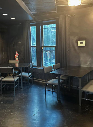 Moody indoor dining area with black-painted walls and floor, simple black tables and gray chairs by tall windows with metal security bars, wall thermostat and fire extinguisher.