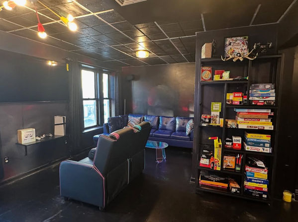 Cozy indoor game lounge with black walls and ceiling, reclining theater seats and a blue sectional by a window, glass coffee table, colorful pendant lights, and a tall shelf packed with board games — casual community game room vibe.