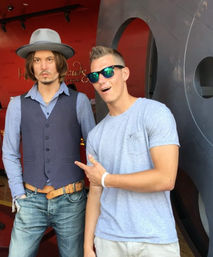 Tourist in sunglasses excitedly points at a lifelike wax figure of a male actor wearing a hat, vest and jeans inside a red-walled wax museum display.