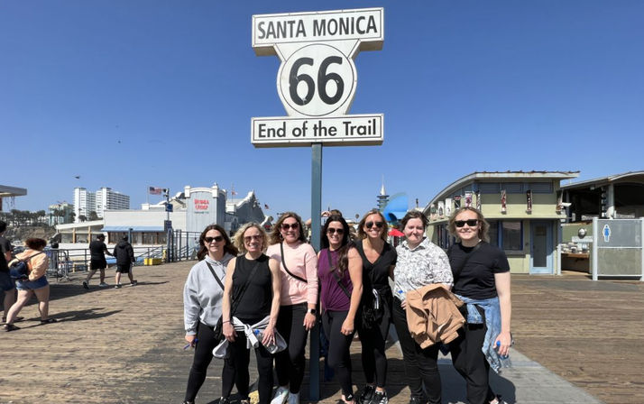 Group of eight women smiling under the Route 66 “Santa Monica – End of the Trail” sign on the Santa Monica Pier boardwalk, bright blue sky and pier attractions in the background.