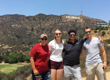 Four friends smiling and posing on a sunny trail with the Hollywood Sign on the Los Angeles hills in the background.