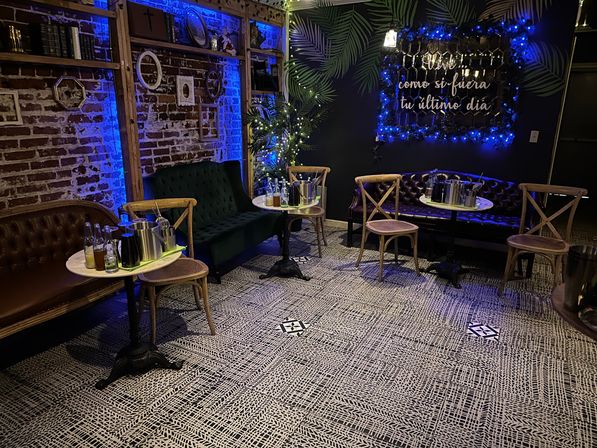 Cozy moody lounge with exposed brick wall and tropical leaf mural, blue string lights and a Spanish neon sign, tufted leather and velvet sofas, wooden bistro chairs and small round tables with bottles and ice buckets.
