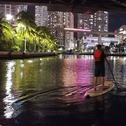 Night paddleboarder gliding on an urban waterfront canal with palm trees and high-rise buildings casting colorful reflections.