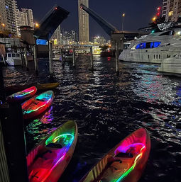 Nighttime urban waterfront with raised drawbridge and skyscraper skyline, luxury yachts docked and colorful LED-lit kayaks in the foreground reflecting neon on rippling water.
