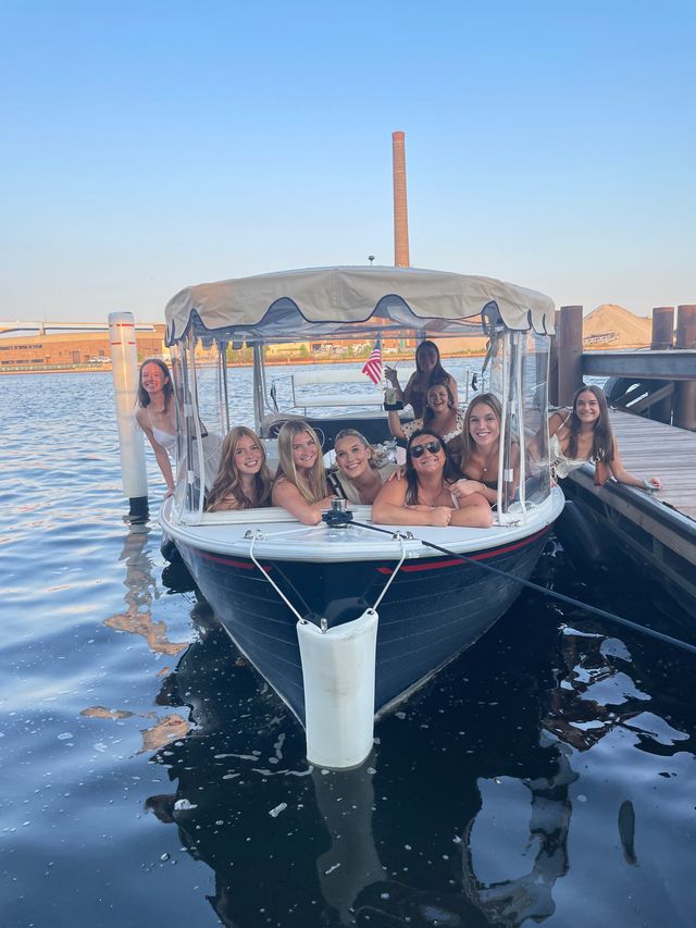 Smiling group of women enjoying a summer boat outing docked at an urban waterfront harbor on a covered leisure boat with a small American flag.