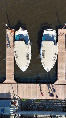 Aerial drone view of two white boats with matching beige canvas covers docked between wooden marina piers on brown river water; people and long shadows stretch across the sunlit dock.