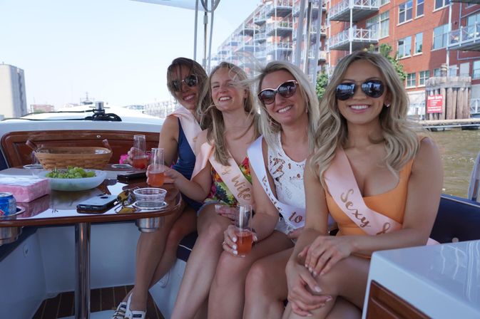 Four smiling women wearing sashes and sunglasses enjoy cocktails on a sunny boat near a city waterfront, with a bowl of grapes on the table — bachelorette party vibe.