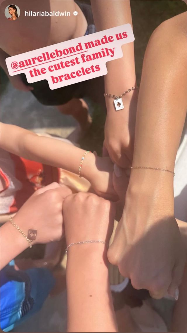 Sunlit close-up of five mixed-age wrists joined in a circle outdoors, each wearing matching delicate chain family bracelets with tiny charms.