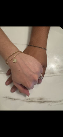 Two hands clasped on a white marble countertop, one wearing stacked gold bracelets with a small clover charm and the other wearing a thin chain bracelet and a sparkling diamond ring.