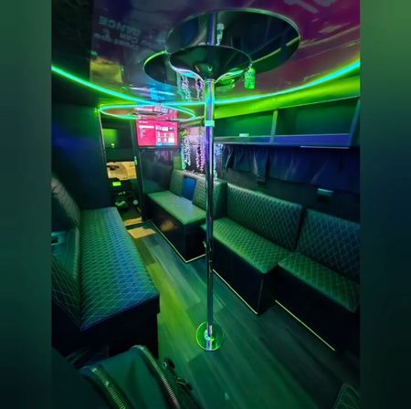 Vibrant neon-lit party bus interior with a chrome dance pole, quilted bench seating along both sides, overhead green-blue LED accent lighting and a wall TV — modern mobile lounge.