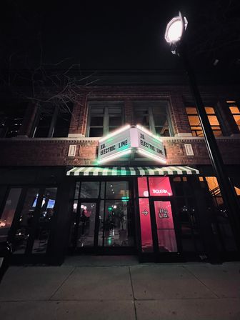 Nighttime downtown storefront — two-story brick building with an illuminated green marquee and striped awning, glass entrance showing pink neon "HOLA" inside and a glowing streetlamp overhead.