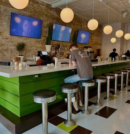 Casual urban cafe/bar interior with a person seated at a white countertop and green bar base, chrome retro stools, exposed brick wall, three wall-mounted TVs and round pendant lights, daytime coffee-and-cocktails vibe.