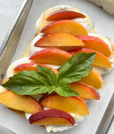 Close-up of ricotta toast topped with sliced ripe peaches, honey drizzle and fresh basil on a parchment-lined baking sheet.