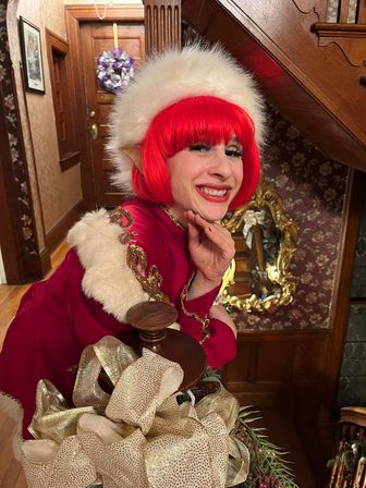 Cheerful holiday elf in a bright red wig and white fur-trimmed hat leaning on a wooden Victorian staircase banister decorated with gold ribbon and greenery.