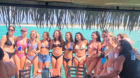 Group of women in colorful bikinis posing under a thatched roof on a boat deck with bright turquoise water and a sandy shoreline — tropical beach boat party photo