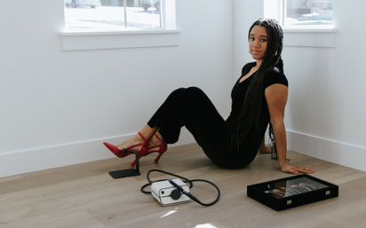 Person with long braids in a black outfit and red heels seated on light wood floor in a bright minimalist corner by windows, glancing at the camera; a jewelry display box and a white portable device with a coiled cable sit nearby.