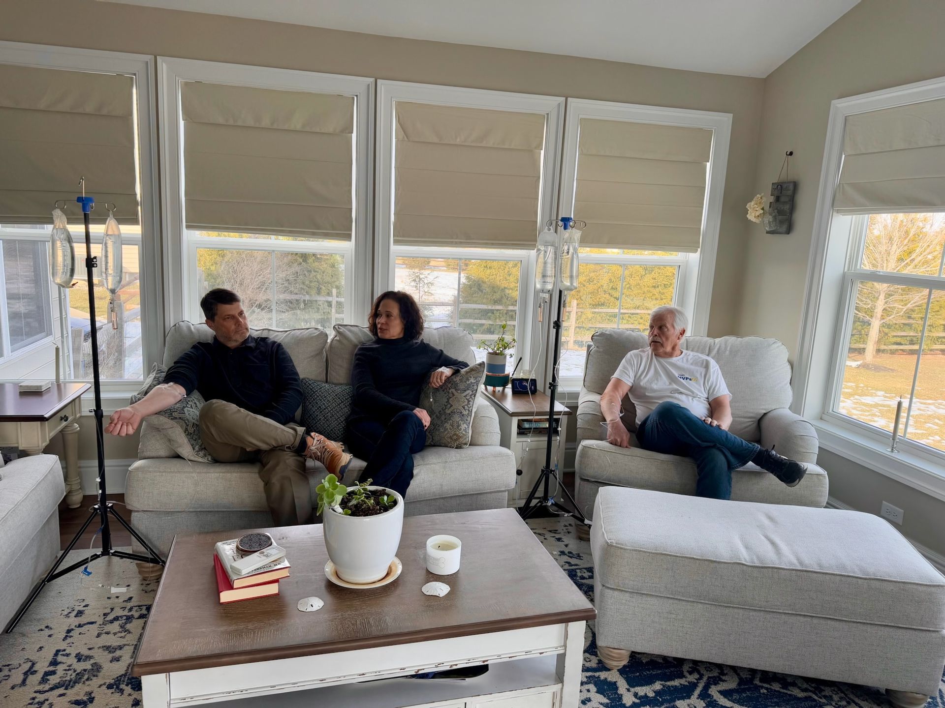 Three adults sitting on light-gray sofas in a sunlit cozy living room with large windows and beige Roman shades; two IV drip stands beside the seating, a wooden coffee table with a potted plant, books and candle, and a view of a suburban yard.