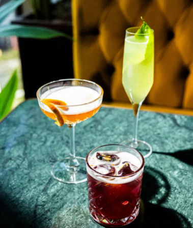 Trio of vibrant cocktails — orange coupe with citrus twist, pale green flute with leaf garnish, and red iced lowball — on a green marble table by a mustard tufted booth in a cozy bar