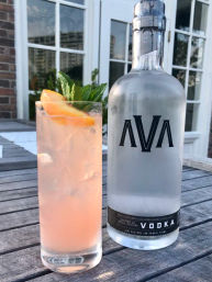 Pink grapefruit vodka cocktail with ice, mint sprig, and grapefruit wedge in a tall glass beside a frosted vodka bottle on a wooden outdoor patio table in front of sunlit windows