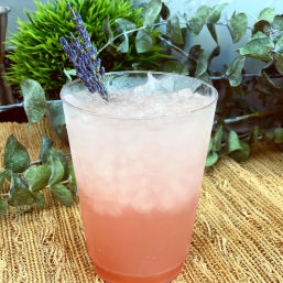 Refreshing pink lavender lemonade in a tall glass filled with crushed ice and garnished with a lavender sprig, photographed on a woven mat with eucalyptus and green plants in the background