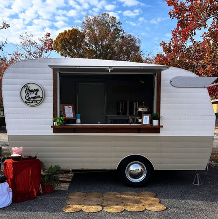 Retro cream-and-beige vintage camper converted into a mobile bar with open serving window, wood counter, small menu sign and potted plants, parked on a leafy autumn street with woven mats in front.