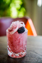 Pink blackberry crushed-ice cocktail in a cut crystal glass, topped with a fresh blackberry and a pale curled rhubarb ribbon, on a wooden table with soft bokeh background