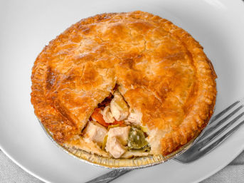 Golden flaky chicken pot pie in a foil tin with a wedge removed to reveal chunks of chicken and mixed vegetables, served on a white plate with a fork