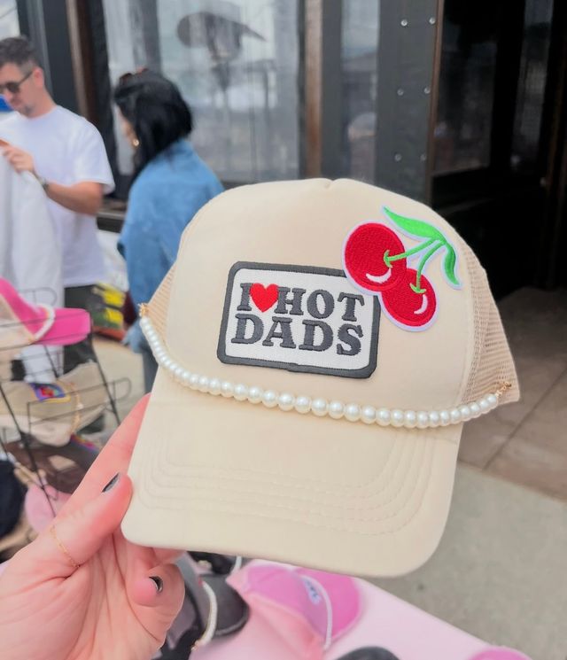 Beige trucker hat with pearl trim, embroidered cherry patch and a sewn label reading "I ♥ HOT DADS," held above a blurred outdoor market stall.