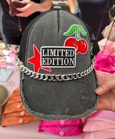 Distressed black trucker hat on a vendor table with a 'LIMITED EDITION' patch, red star and cherry patches, and a silver chain trim across the frayed brim.