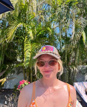 Smiling woman in sunglasses and a colorful “Have a Nice Day” cap wearing an orange patterned swimsuit, poolside with tropical palm trees and bright flowers on a sunny day