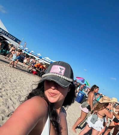 Selfie of a woman in a pearl-trimmed cap at a crowded sunny beach festival — sandy shoreline, vendor tents and colorful umbrellas, people in swimwear under a bright blue sky.