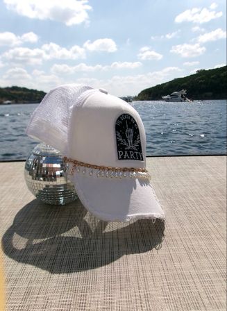 Party-ready white trucker cap with pearl chain draped over a small disco ball on a boat dock, sparkling lake water and a yacht under a sunny summer sky