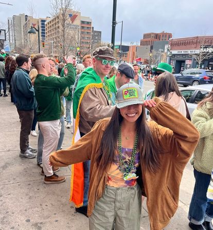 Smiling woman in a trucker hat and green beads poses on a busy downtown sidewalk with a festive crowd wearing green and shamrock accessories for St. Patrick’s Day celebrations.